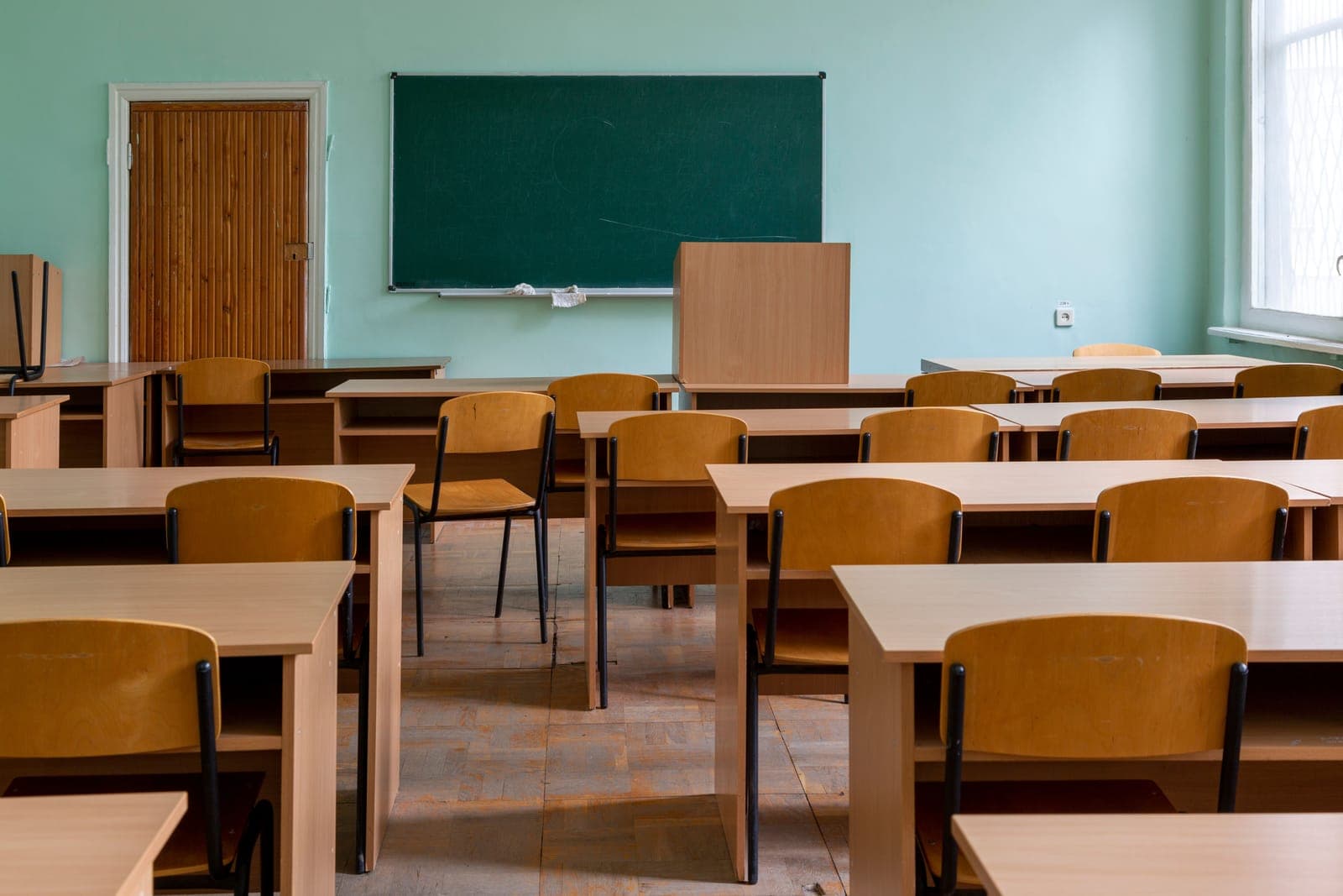 An empty classroom with light desks, wooden chairs and a green chalkboard on a mint-green wall.